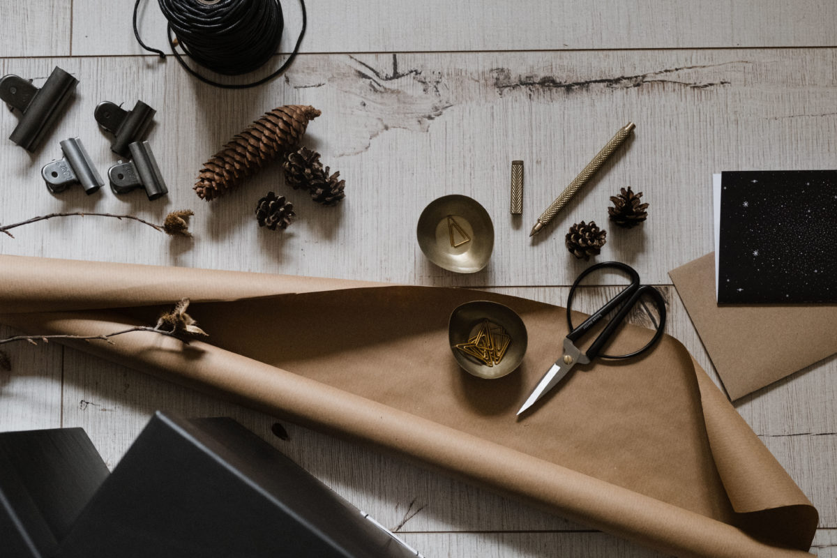 Weihnachtsstilleben mit Geschenkpapier, Schere, Band, Tannenzapfen, Geschenkkarte, Schälchen, Stift und Klammern